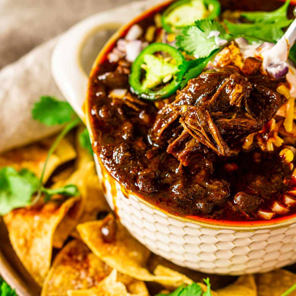 A spoon scooping out some short rib chili in a cream-colored bowl.