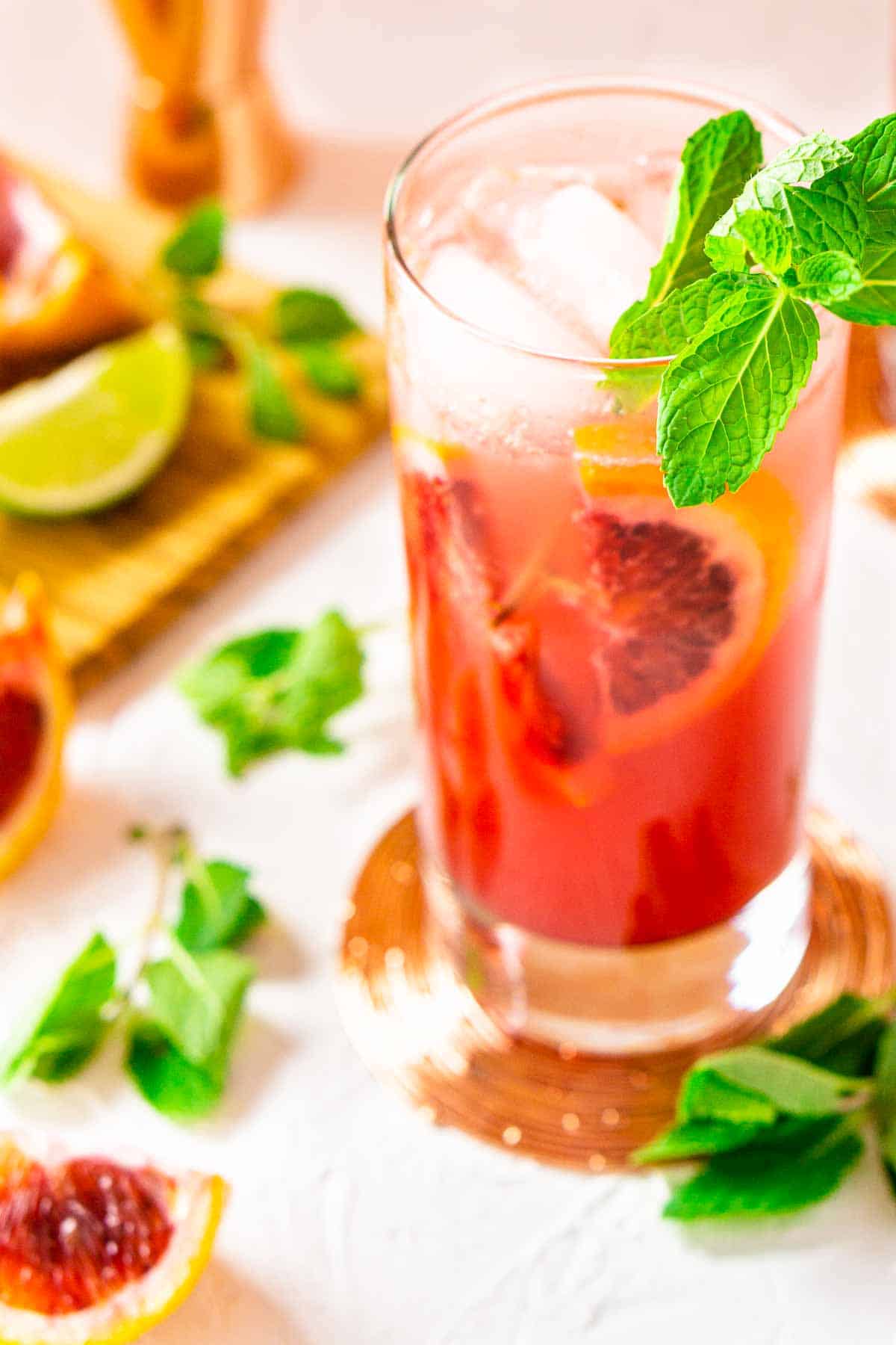 A blood orange mojito on a copper coaster with fruit around it.