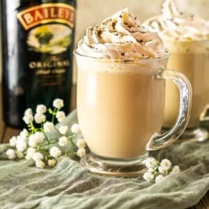 A Baileys latte on a green clothe with a bottle of Irish cream behind it.
