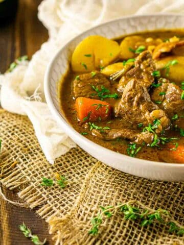 A bowl of Guinness lamb stew on burlap with herbs and beer to the side.