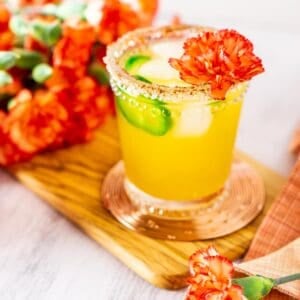 A jalapeno-mango margarita on a wooden board with a colorful napkin and flowers around it.