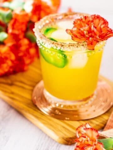 A jalapeno-mango margarita on a wooden board with a colorful napkin and flowers around it.
