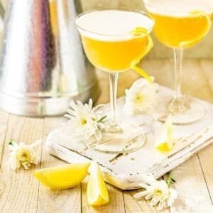 Two rum sour cocktails on a white wooden board with flowers and lemon slices around them.