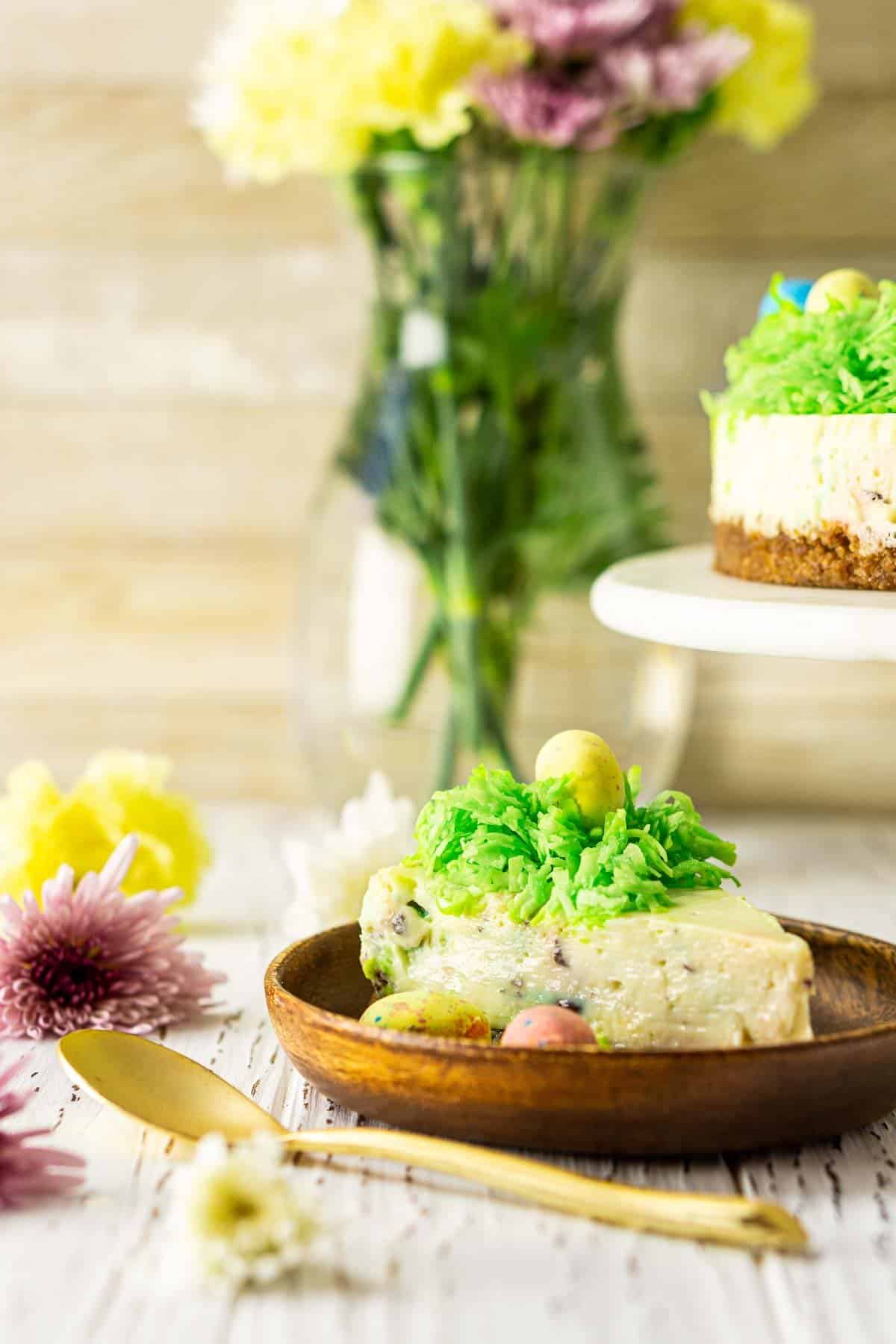 A slice of Easter egg cheesecake on a wooden plate with spring flowers around it and a gold spoon in front.