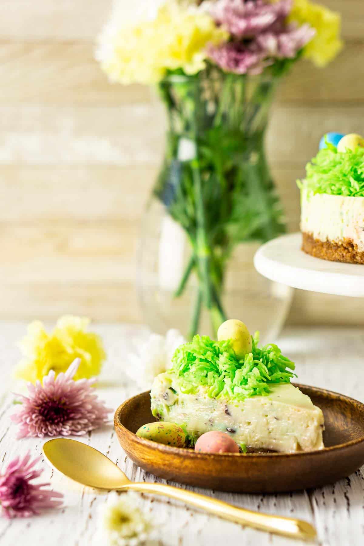 The Easter egg cheesecake with a gold spoon on a wooden plate with flowers to the side.