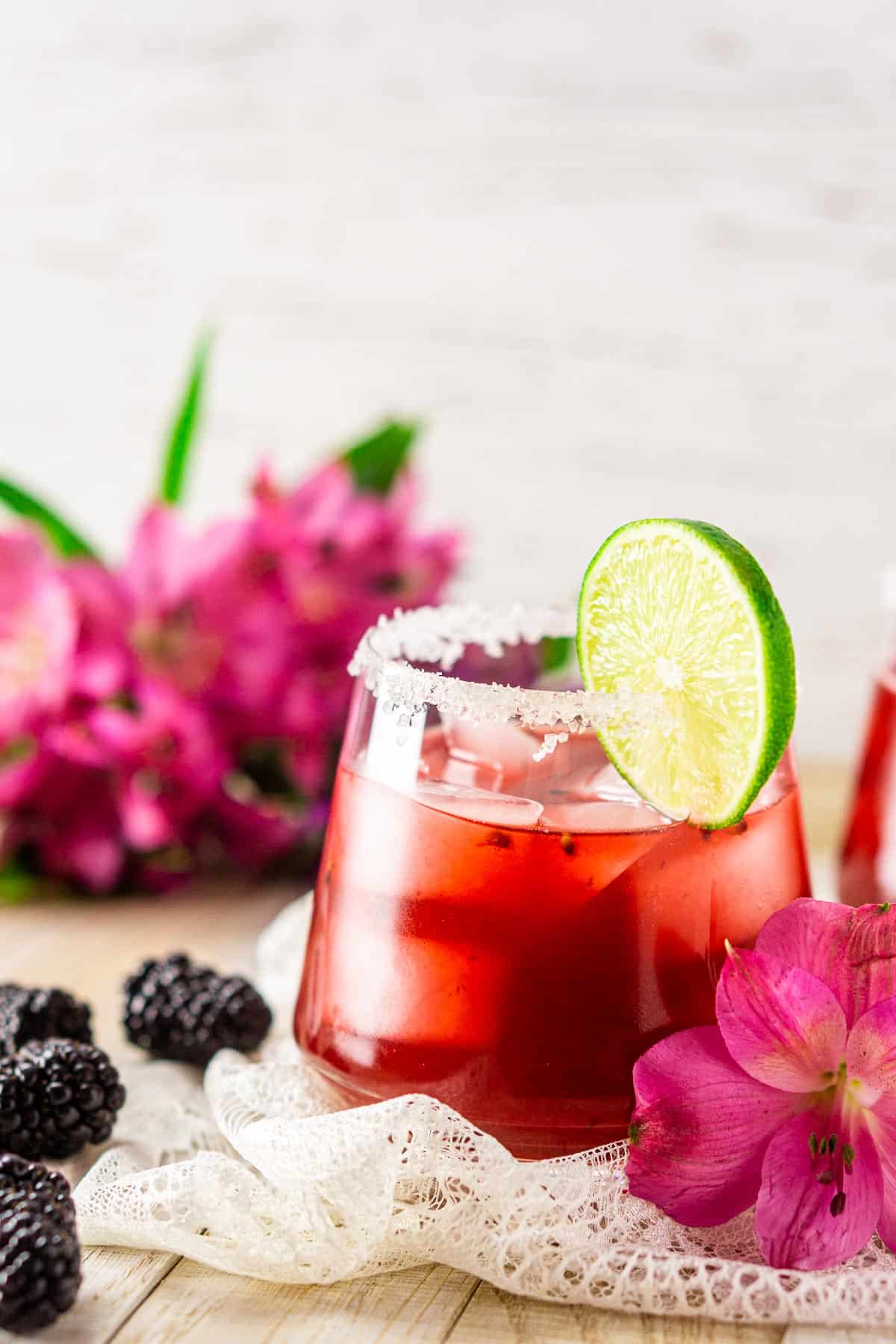 A blackberry margarita with pink flowers on white lace with fruit to the side.