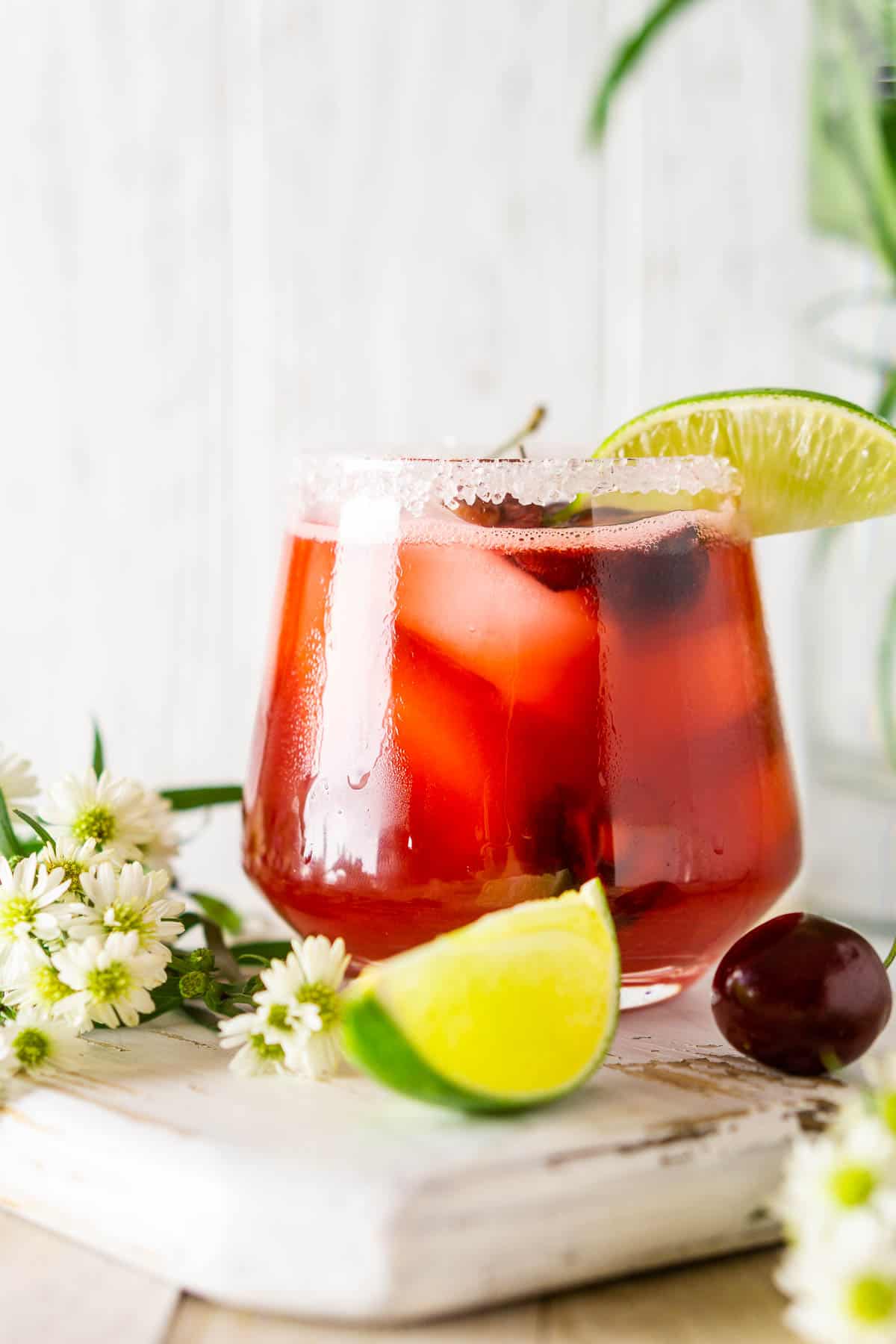 A cherry margarita with white flowers and limes and cherries around it.