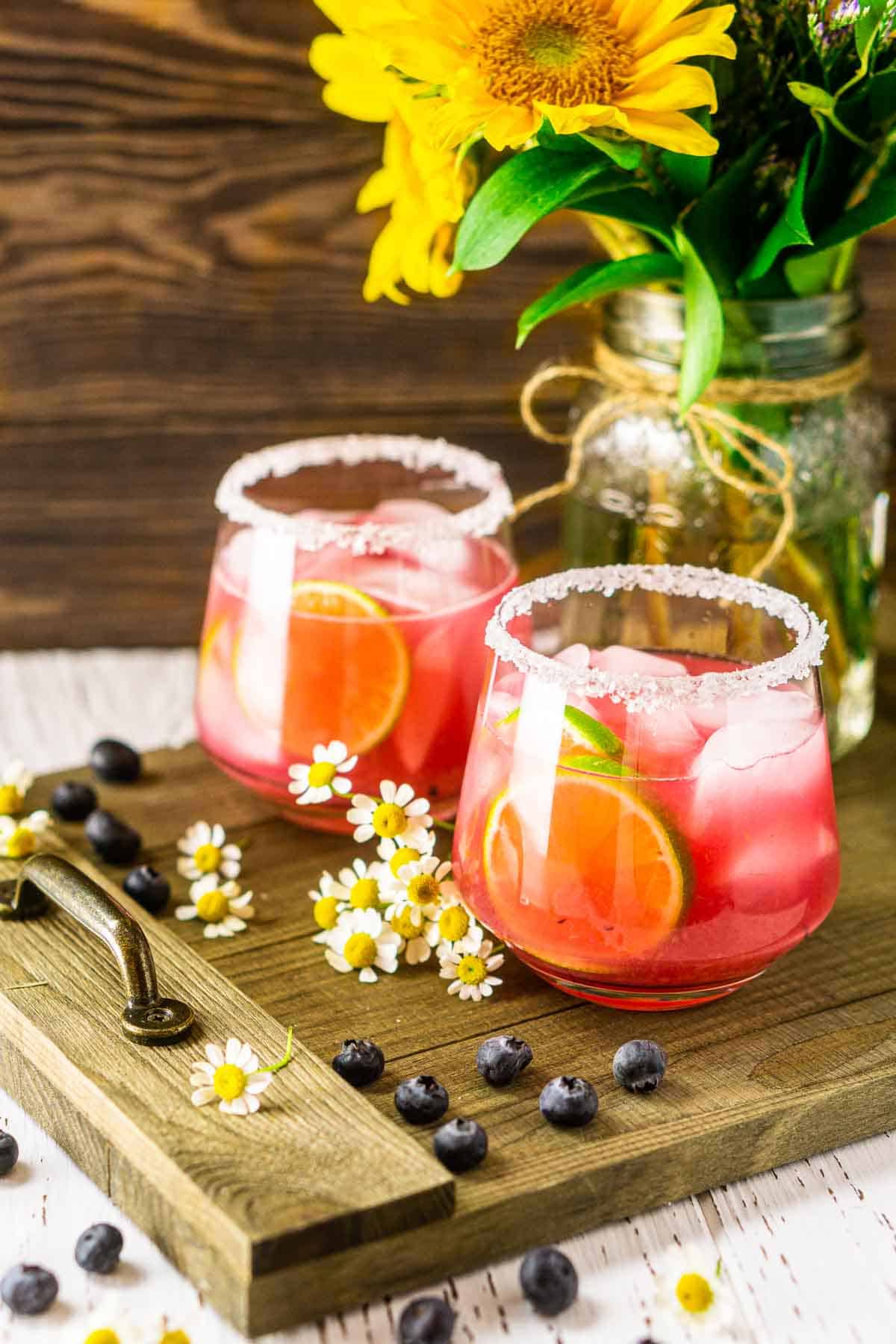 Two blueberry margaritas on a wooden serving tray with fresh berries all around them.