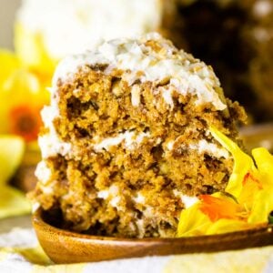 A slice of homemade carrot cake on a wooden plate on top of a yellow and white napkin with a yellow flower to the side.