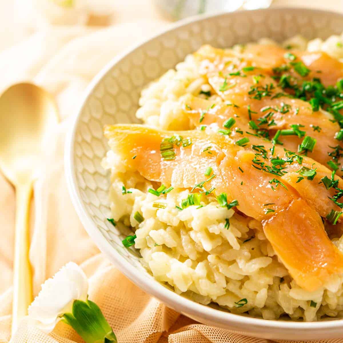 A bowl of smoked salmon risotto with white flowers and a gold spoon to the left.