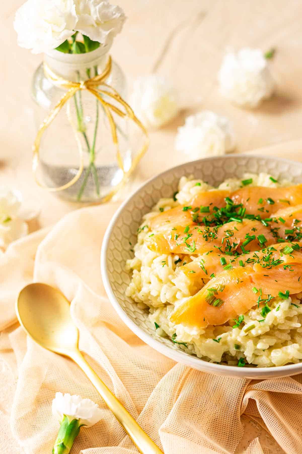 Looking down on a bowl of smoked salmon risotto with a small vase of white flowers and a gold spoon on the left.