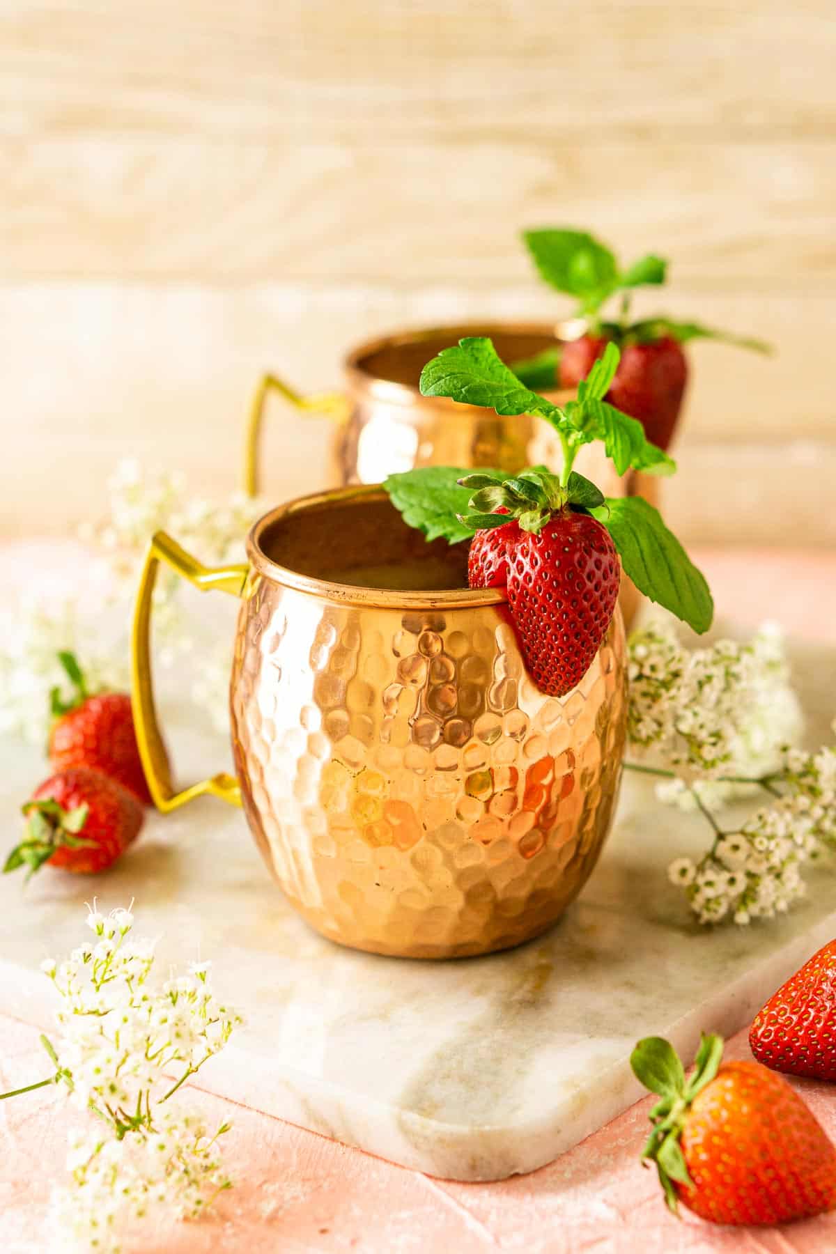 A close-up of a minted strawberry Moscow mule surrounded by flowers and berries.