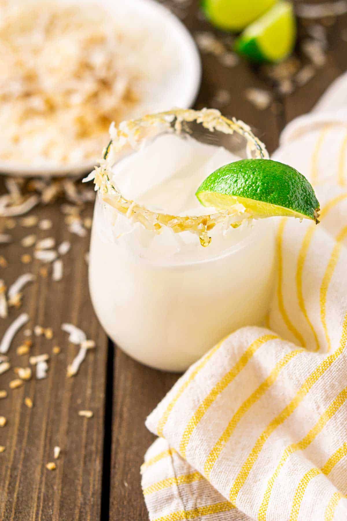 A coconut margarita with a yellow and white napkin to the right and a plate of coconut behind it.