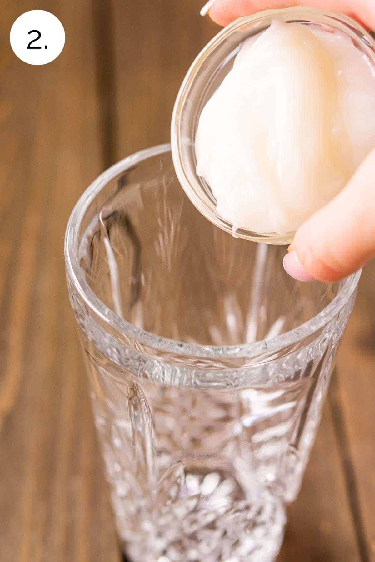 Pouring cream of coconut into a clear crystal cocktail shaker on a wooden board.