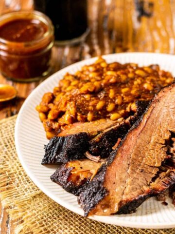 The pellet grill brisket on a white plate with a side of baked beans and BBQ sauce and a beer in the background.