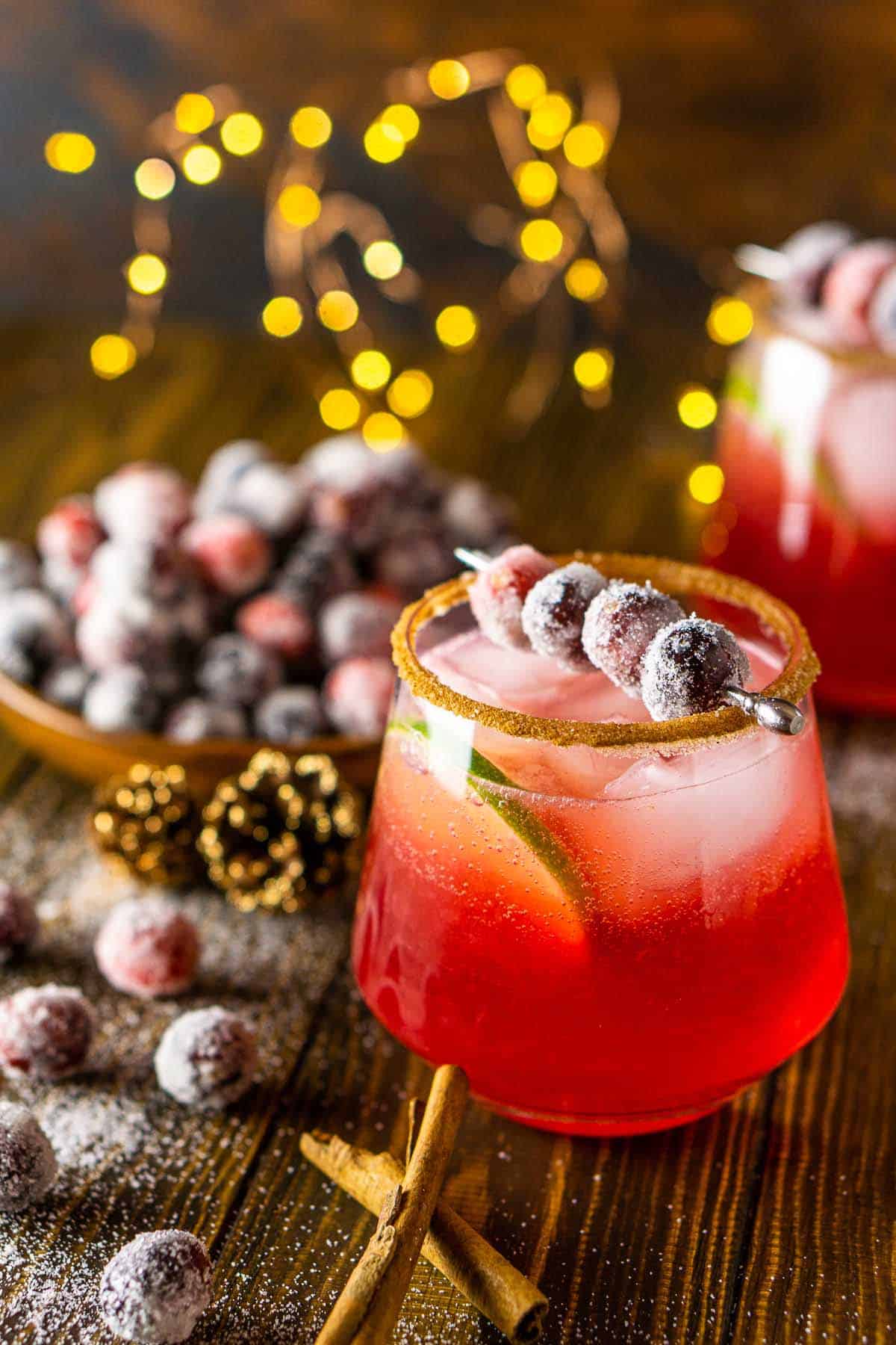 Two cranberry-ginger margaritas on a wooden board with cinnamon sticks and sugared cranberries.
