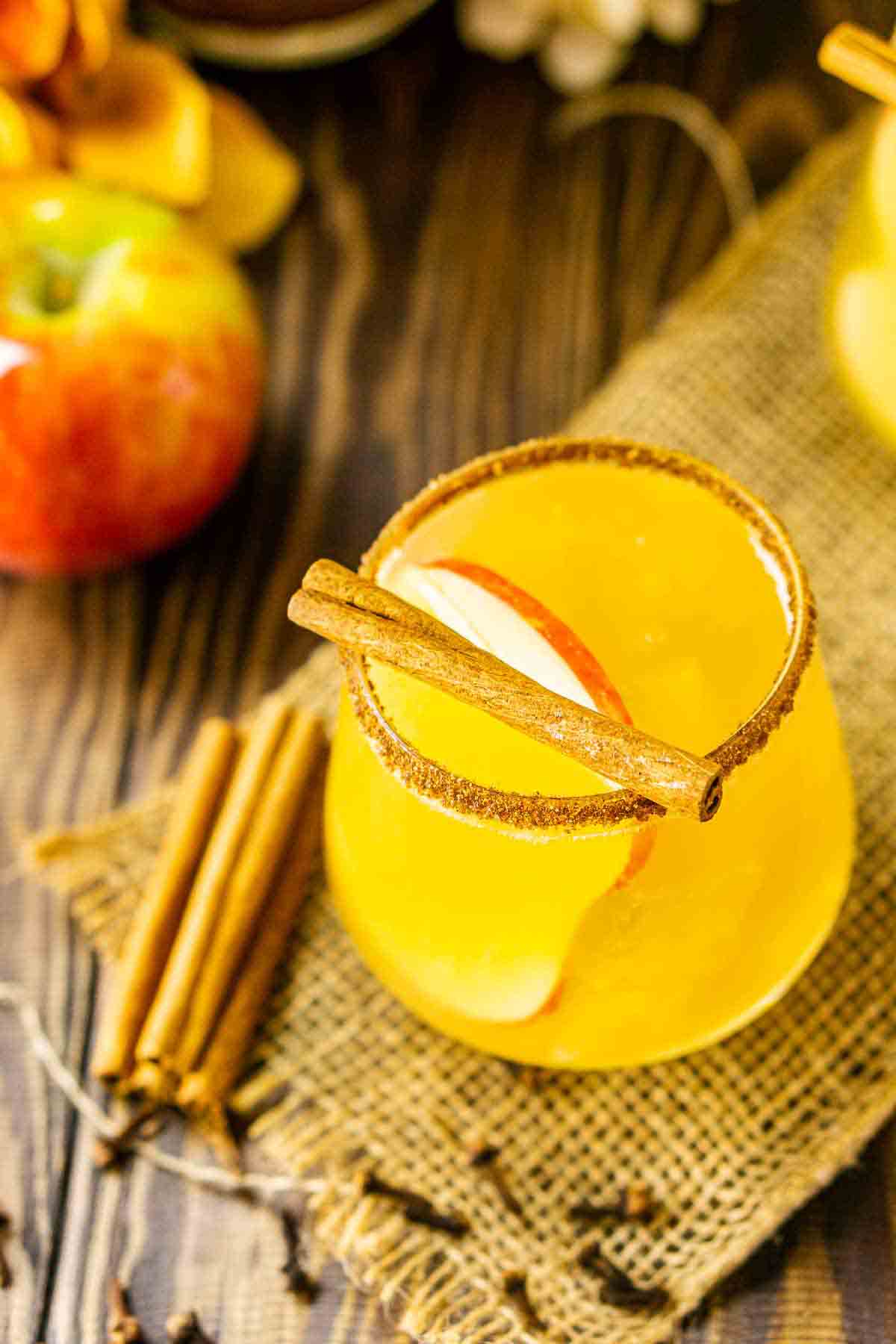 A sparkling apple cider margarita on burlap with cloves and apples around it.