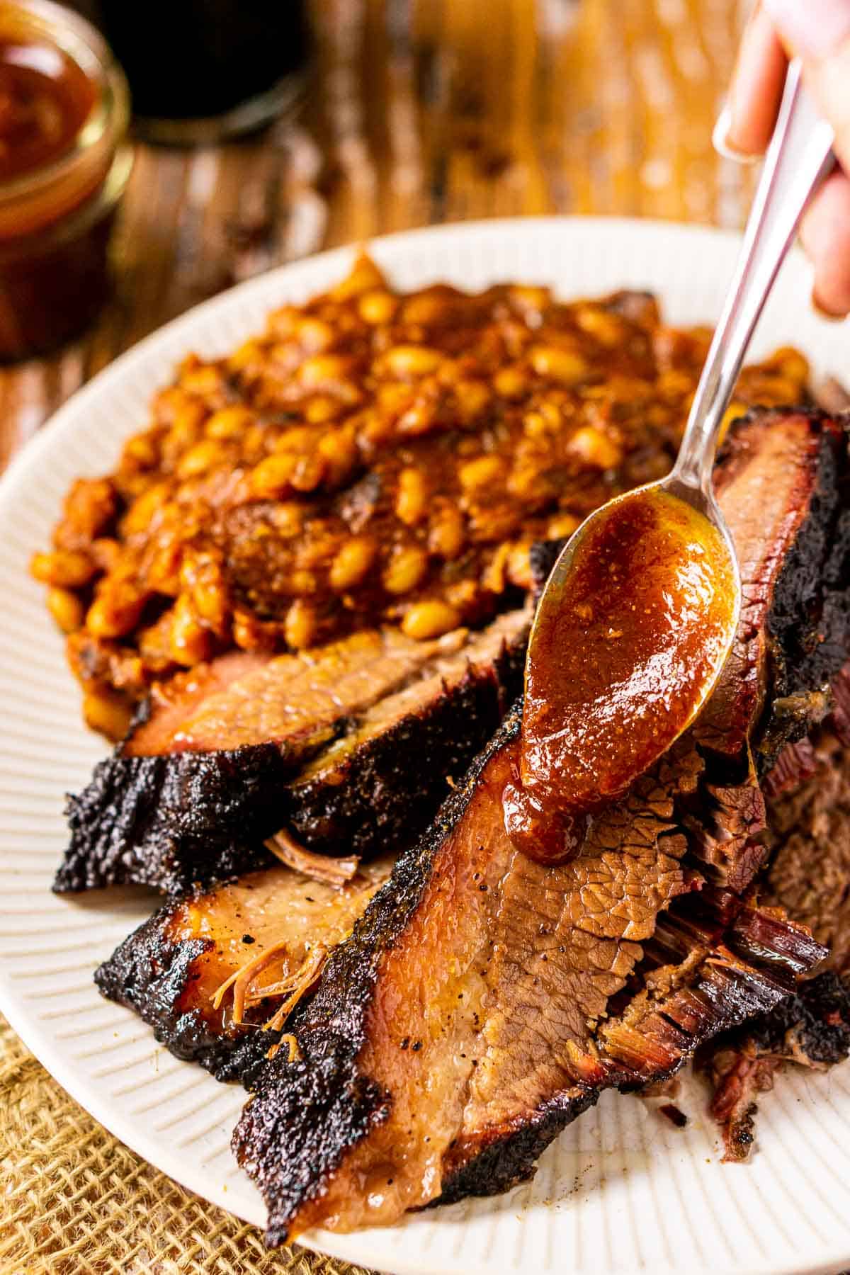 Spooning BBQ sauce onto the slices of brisket on a white plate.