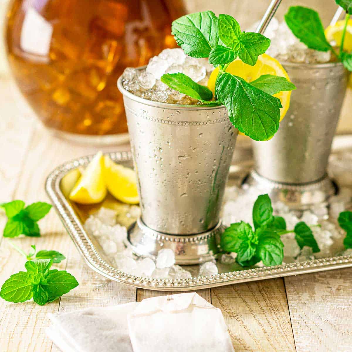 Two sweet tea mint juleps on a silver tray with crushed ice, mint and tea bags around them.