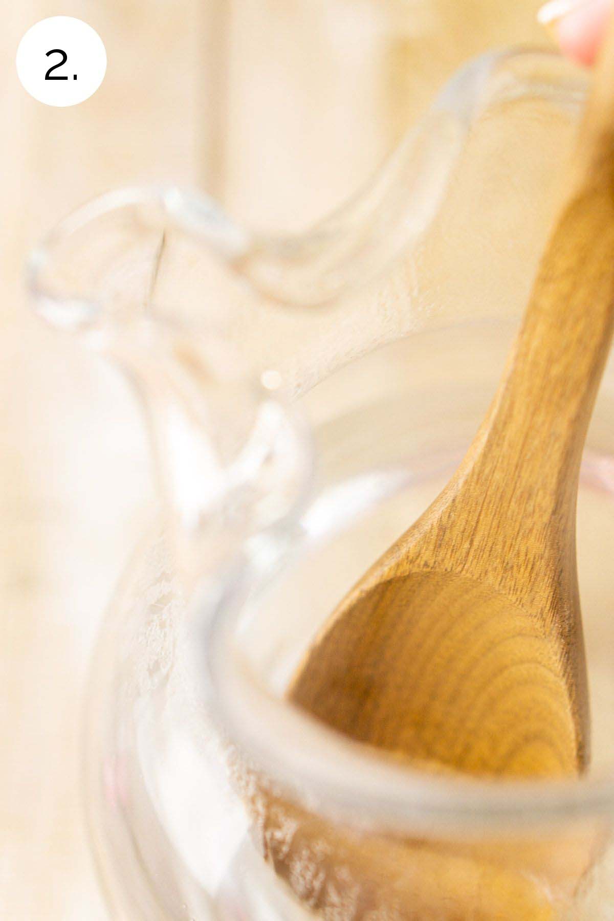 Stirring the sugar and hot water with a wooden spoon in a clear pitcher.