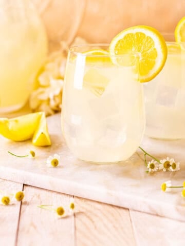 Looking straight on to two tequila lemonade cocktails with a pitcher behind them and white flowers all around.