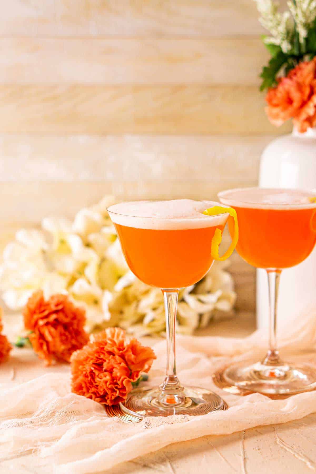 Looking straight on to an Aperol sour with a white vase next to it with orange and white flowers.
