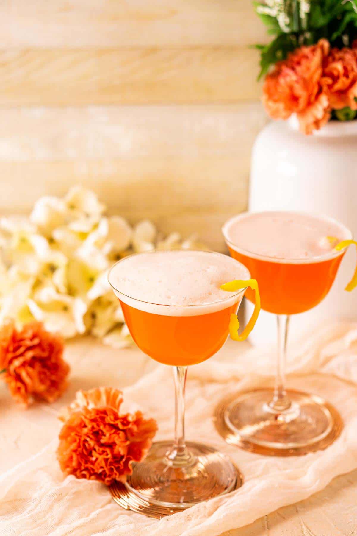 Looking down on two Aperol sour cocktails with an orange flower to the side and a bundle of white flowers in the background.