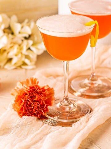 Two Aperol sour cocktails on a pink clothe with orange flowers around them.