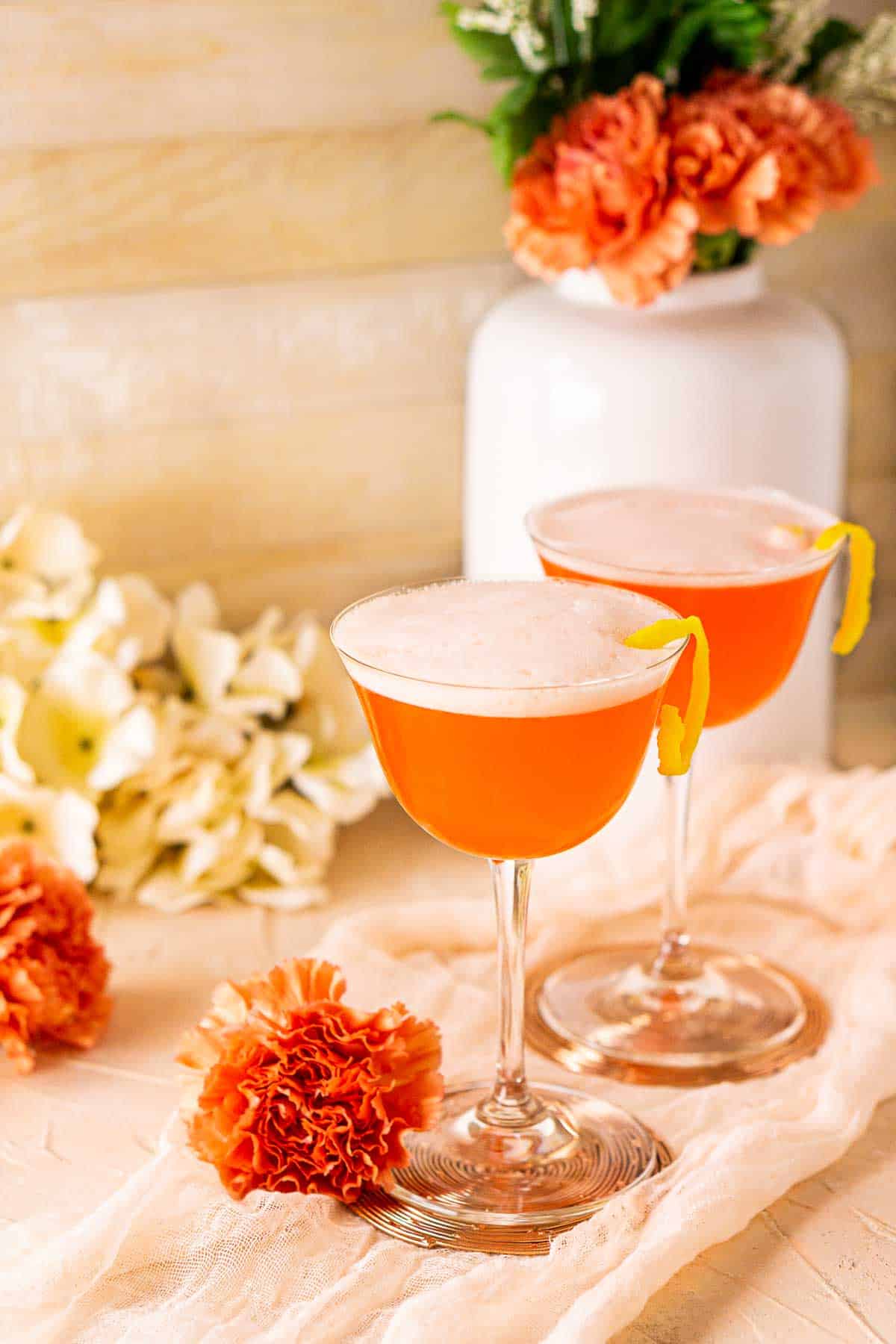 Two Aperol sour cocktails on a pink clothe with orange flowers around them.