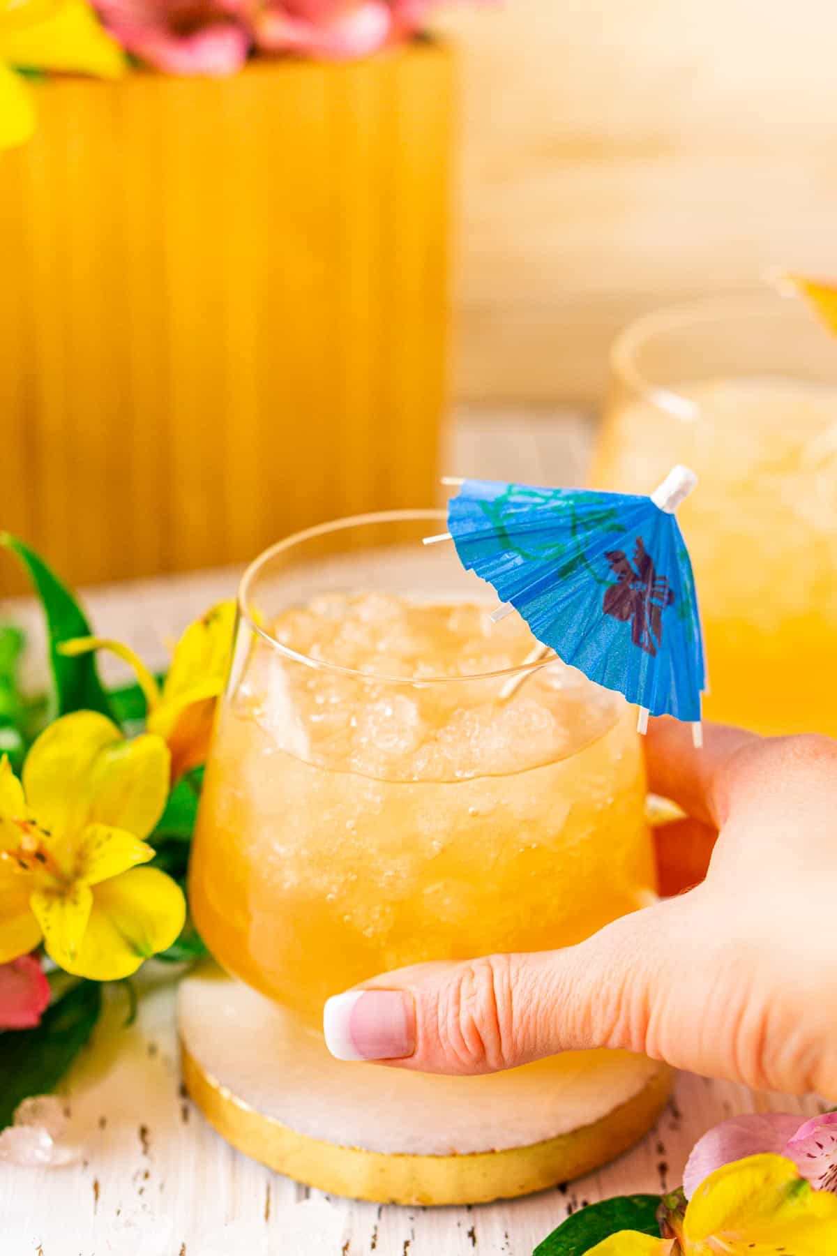 A hand grabbing a glass of the Barbados rum punch on a white coaster with flowers around it.