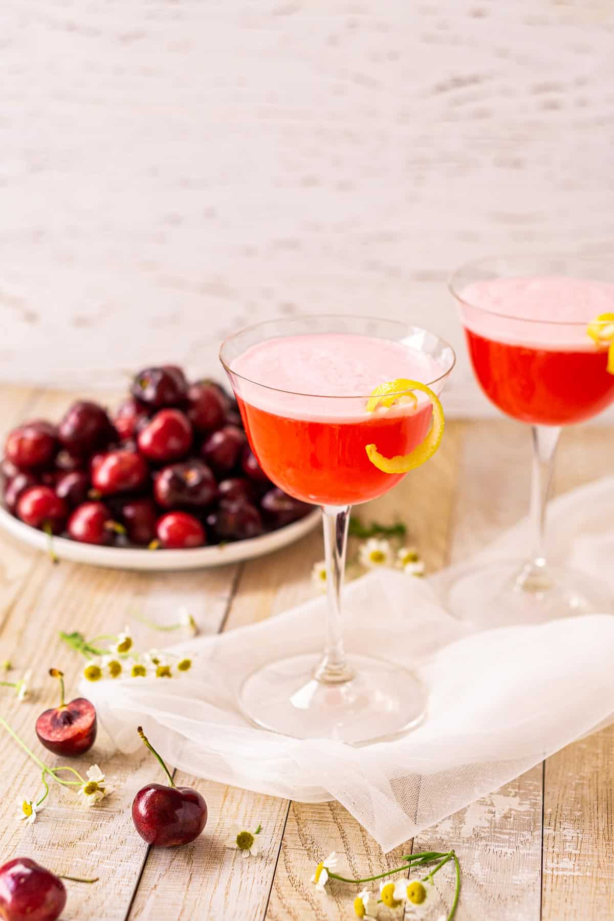 A side view of the cherry vodka sour on a lightly colored wooden board with cherries and flowers.