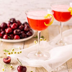 Два кислых коктейля с вишневой водкой на белой сетке с белыми цветами и вишнями вокруг них.