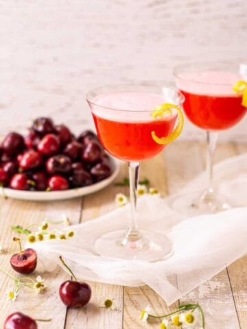 Two cherry vodka sour cocktails on white netting with white flowers and cherries around them.