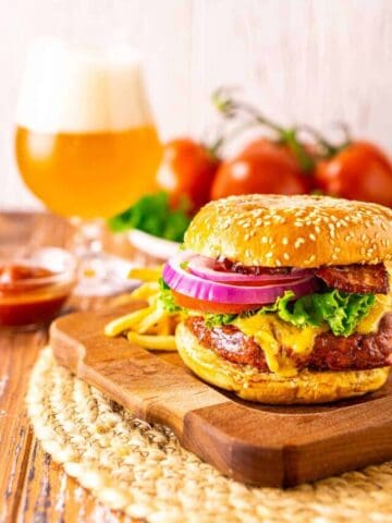 A smoked burger on a wooden surface with fries and a beer in the background.