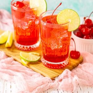 Two Dirty Shirley cocktails on a wooden board with a bowl of cherries to the side and slices of citrus on the other side.