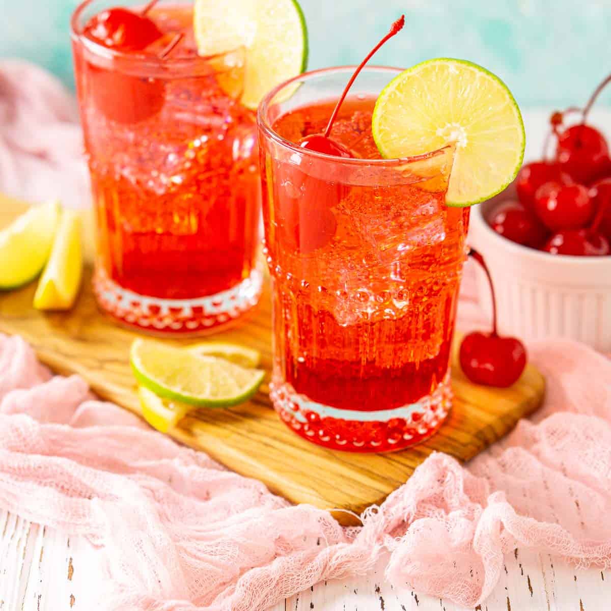 Two Dirty Shirley cocktails on a wooden board with cherries and citrus wedges around them.