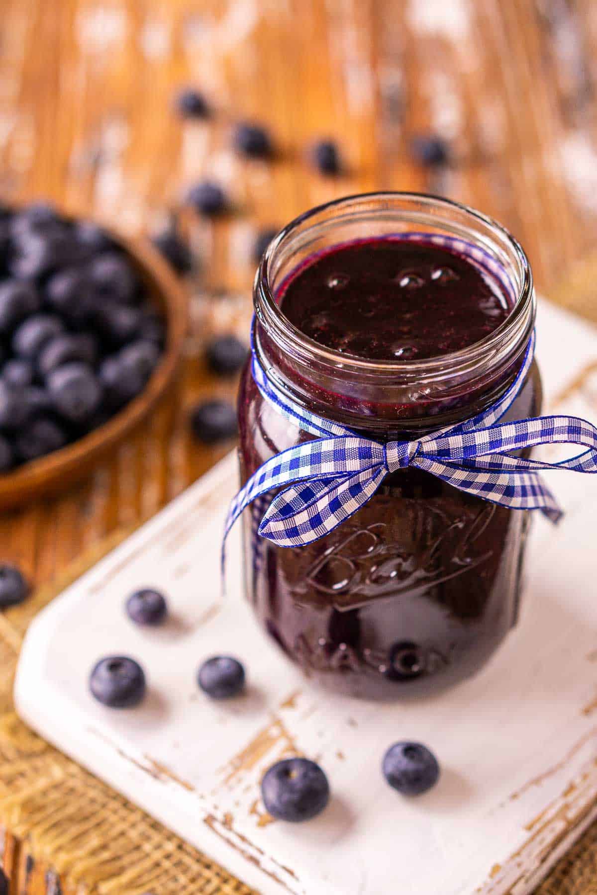 A jar of blueberry BBQ sauce with berries all around it and a plate of berries in the background.