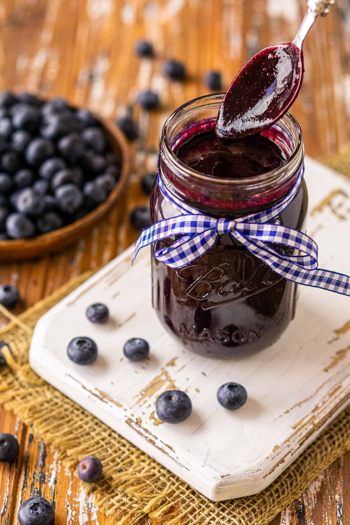 A spoon dripping off BBQ sauce over the jar with blueberries all around it.