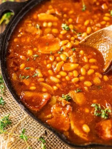 A cast-iron skillet filled with smoked baked beans on burlap with fresh thyme to the side.