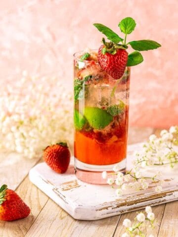 A strawberry mojito on a white wooden board with flowers and fruit around it.