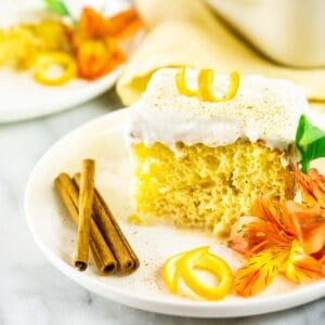 A piece of the spiced orange tres leches cake on a white plate with an orange flower and cinnamon sticks.