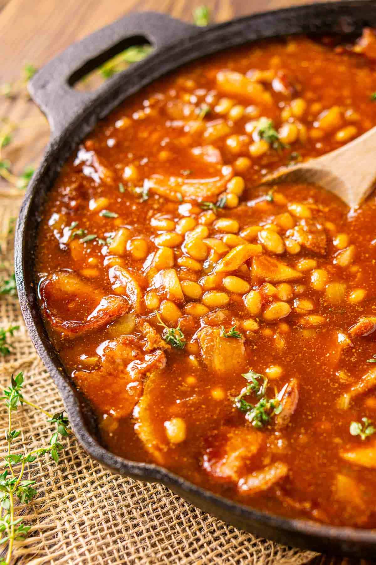 A side view of the smoked baked beans with a spoon in the skillet on burlap.