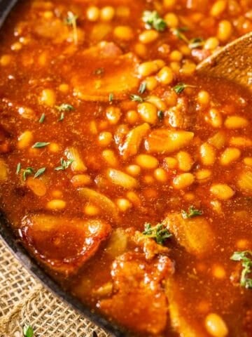 A cast-iron skillet filled with smoked baked beans on burlap with fresh thyme to the side.