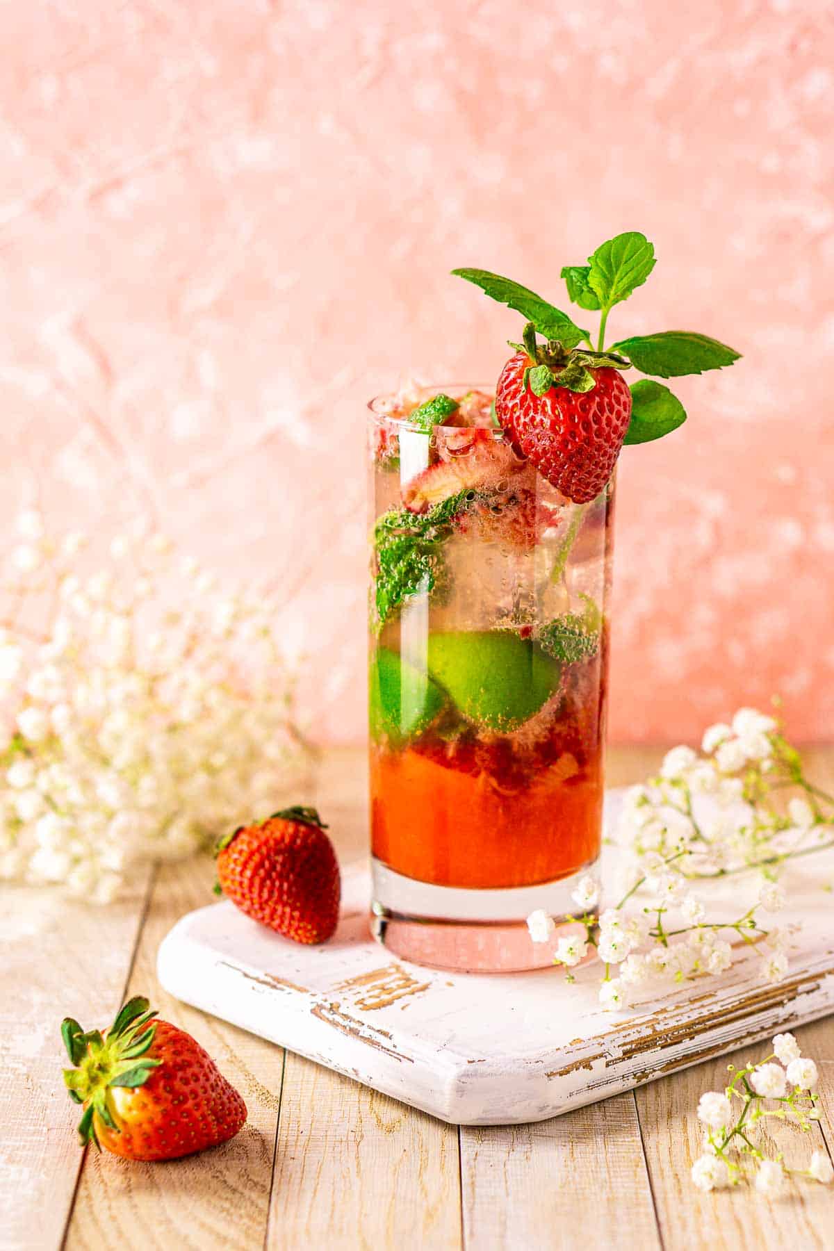 Looking at the strawberry mojito from the side on a white tray with berries and flowers around it.