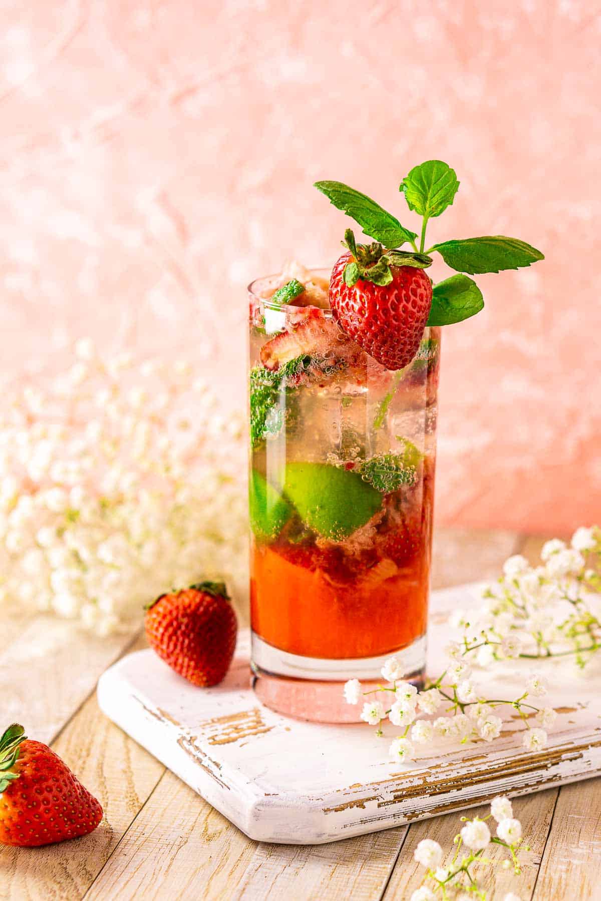 A strawberry mojito on a white wooden board with flowers and fruit around it.