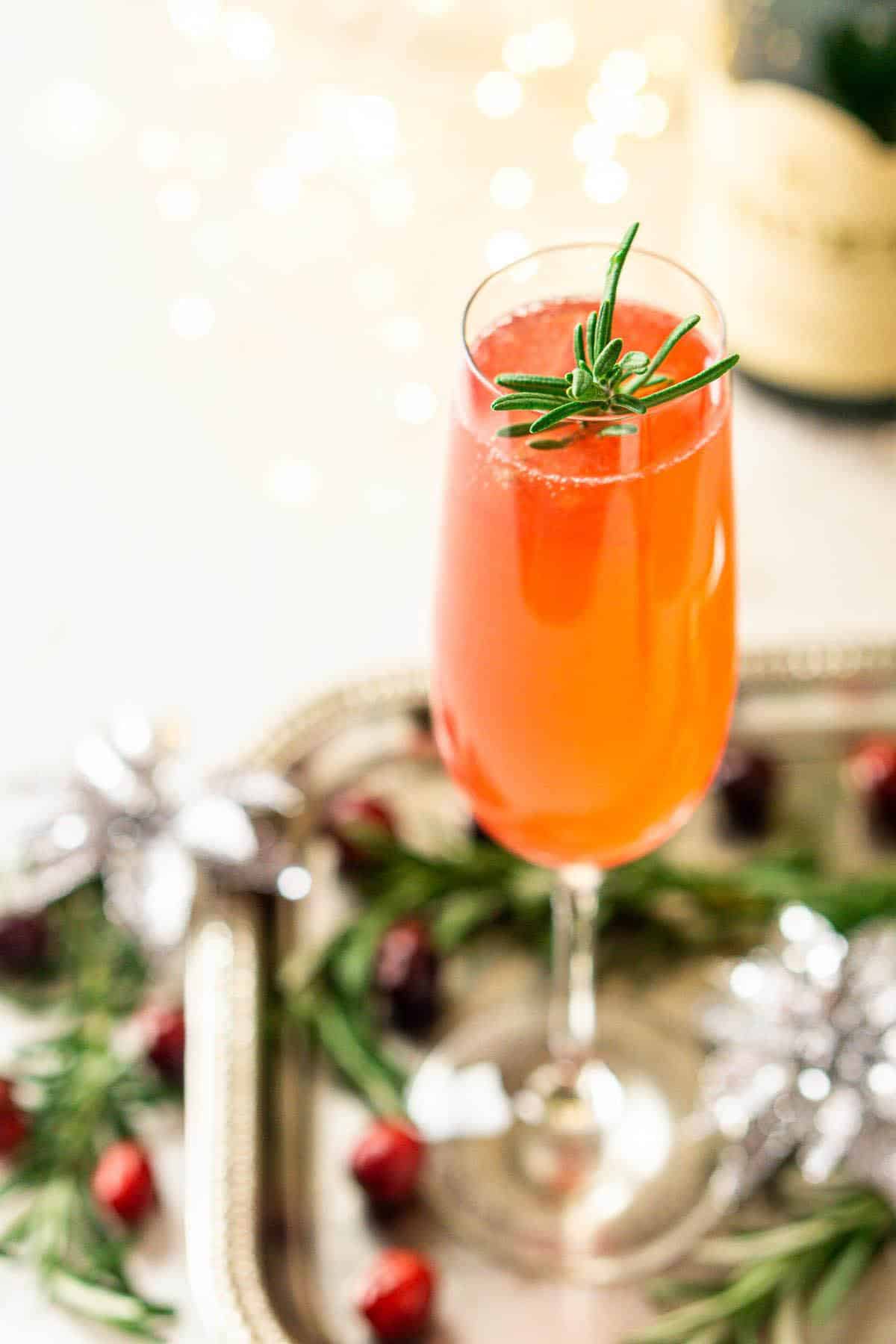 A cranberry French 75 cocktail on a silver tray with fresh rosemary sprigs around it.