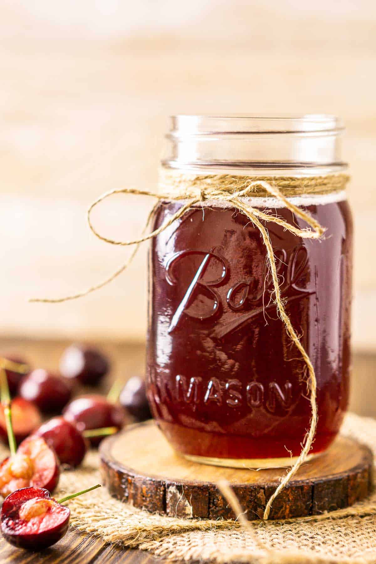Looking straight on to a mason jar of cherry simple syrup on a wooden coaster with burlap underneath.