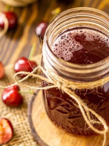 A mason jar on a wooden coaster filled with the cherry simple syrup with fresh cherries all around it on a wooden surface.