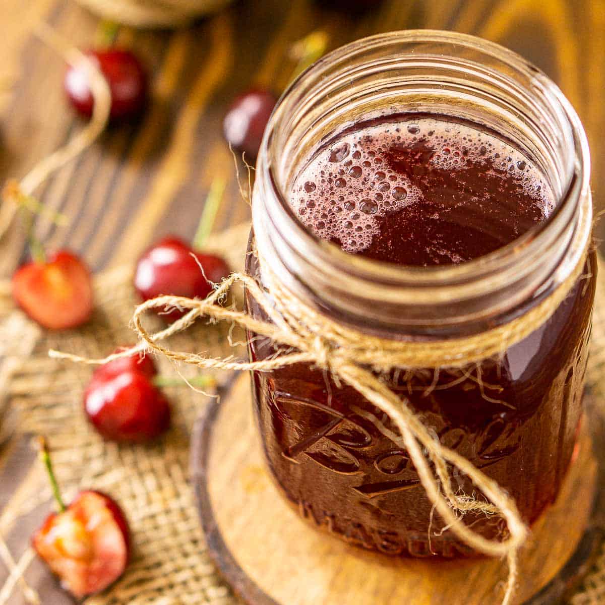 A jar of cherry simple syrup on a wooden coaster with real cherries to the left.