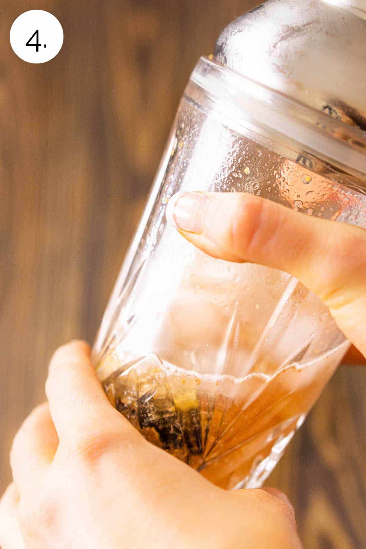 Two hands shaking the cocktail shaker to mix the drink against a wooden surface.